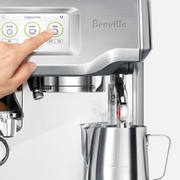 Breville | Barista Touch espresso machine with touchscreen, showing a hand selecting Milk while frothing milk, alongside a loaded portafilter, at Athens Cooks.