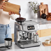 Breville | The Oracle Jet, Black Truffle espresso machine with beans being poured into the hopper, surrounded by a barista setup including a knock-box and milk pitcher.