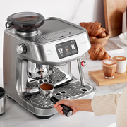 Breville | The Oracle Jet, Black Truffle espresso machine with touchscreen and grinder, on a marble countertop, portafilter held, croissants and latte art drinks in background.