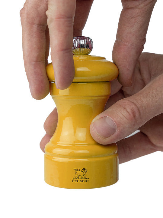 Peugeot | Bistro Pepper Mill, Saffron, shown in use as hands turn its top, highlighting its elegant, glossy design crafted from sustainably sourced French wood.