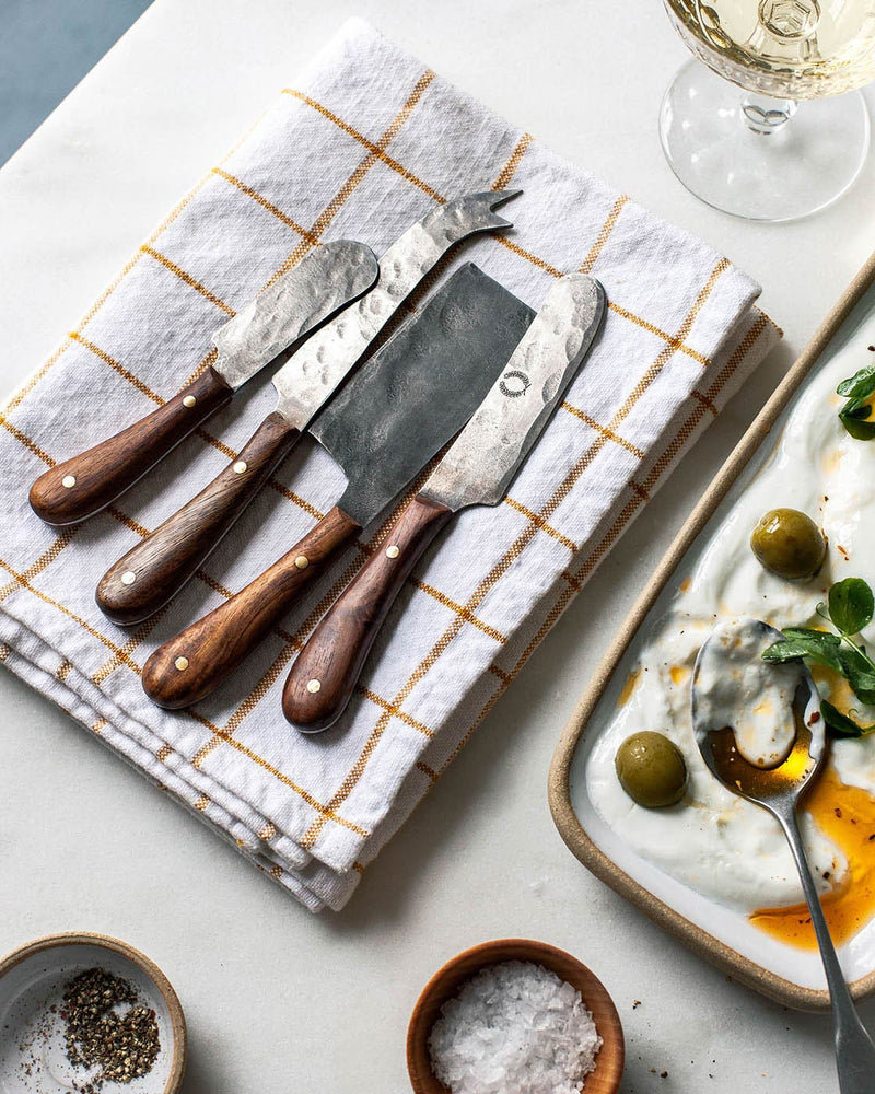 Farmhouse Pottery | Artisan Forged Cheese Knives displayed on a tea towel, alongside a dish of cheese with olives and microgreens, perfect for elegant entertaining.