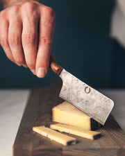 Hand slicing cheese with Farmhouse Pottery | Artisan Forged Cheese Knife, displaying its hammered finish on a wooden board.