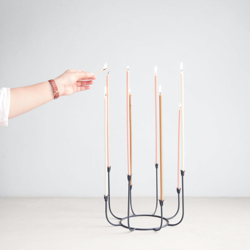 The Floral Society | Beacon Long-Stem Candelabra, Dune: Black metal candelabra with eight pastel tapers, one being lit by hand, showcasing handcrafted elegance from Athens Cooks.