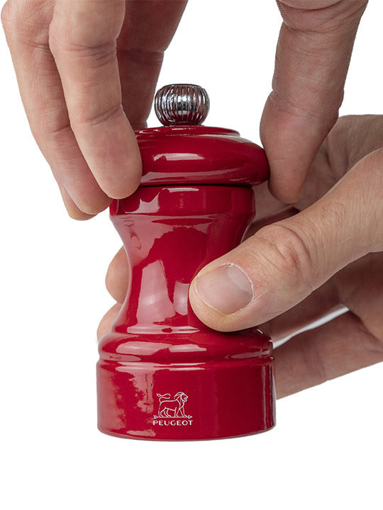 Peugeot | Bistro Pepper Mill, Passion Red, being turned by hand; features a classic design with a metal adjustment knob and embossed lion logo near the base.
