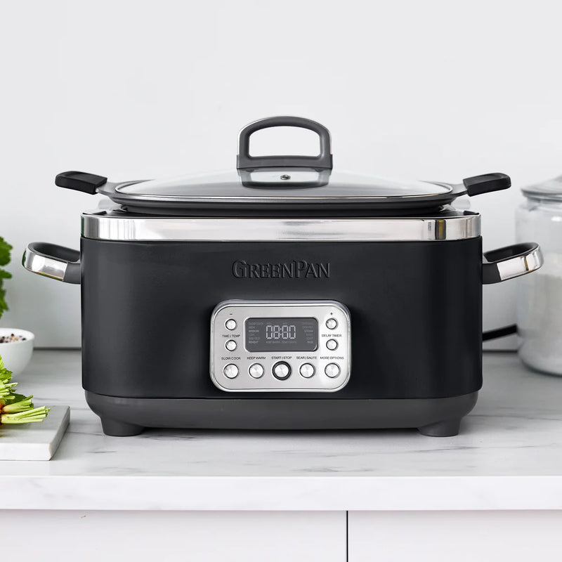 GreenPan Elite 14-in-1 6-Quart Slow Cooker with digital control panel and glass lid, displayed on a kitchen counter next to cooking ingredients.