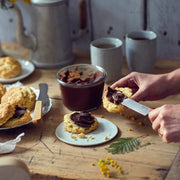 Person spreading chocolate on a scone with Opinel | Brunch Knives, Tangerine, featuring a rounded tip and micro-serrated section, amidst a rustic breakfast setting.