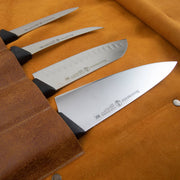 Messermeister Vintage Leather 7 Pocket Knife Roll, Amber, showcasing four professional stainless-steel knives with black handles, partially sheathed in a brown leather roll.