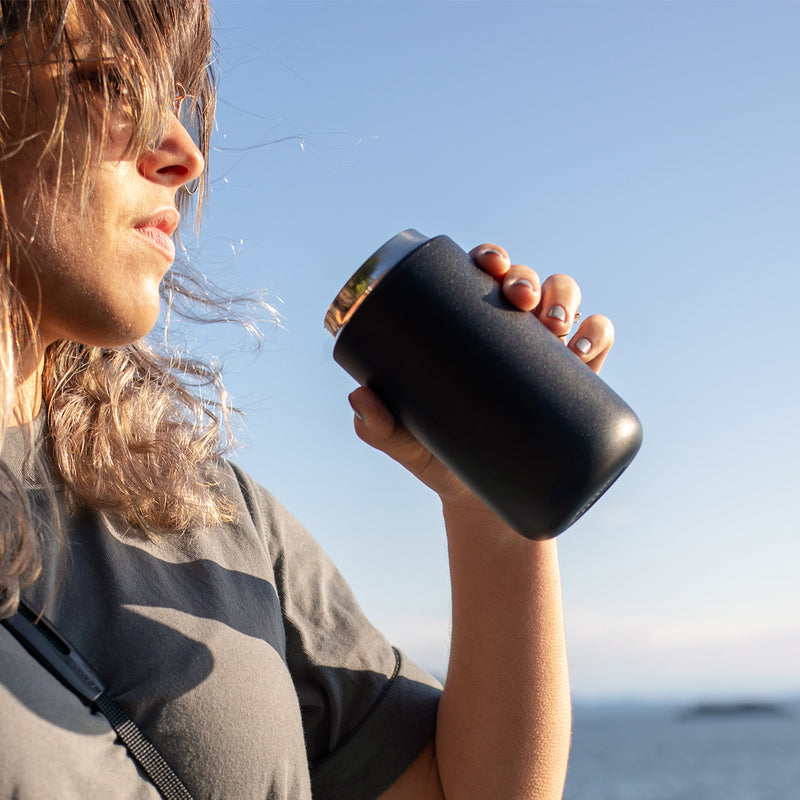 A person enjoys a drink from the FELLOW | Carter Move Mug, Matte Black / 16 oz, showcasing its portability and spill-proof design in an outdoor setting.