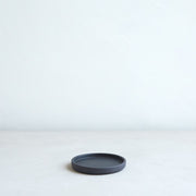 The Floral Society | Daily Plate, Sand: A minimalist black ceramic saucer ideal for candles, jewelry, or desserts, showcased on a light surface against a textured white wall.