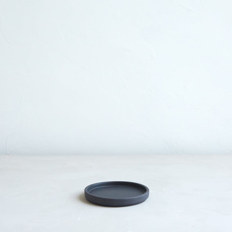 The Floral Society | Daily Plate, Sand: A minimalist black ceramic saucer ideal for candles, jewelry, or desserts, showcased on a light surface against a textured white wall.