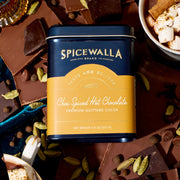 Spicewalla | Chai Spiced Hot Chocolate tin surrounded by broken chocolate pieces and whole spices, with a cup of marshmallow-topped hot chocolate in the corner.