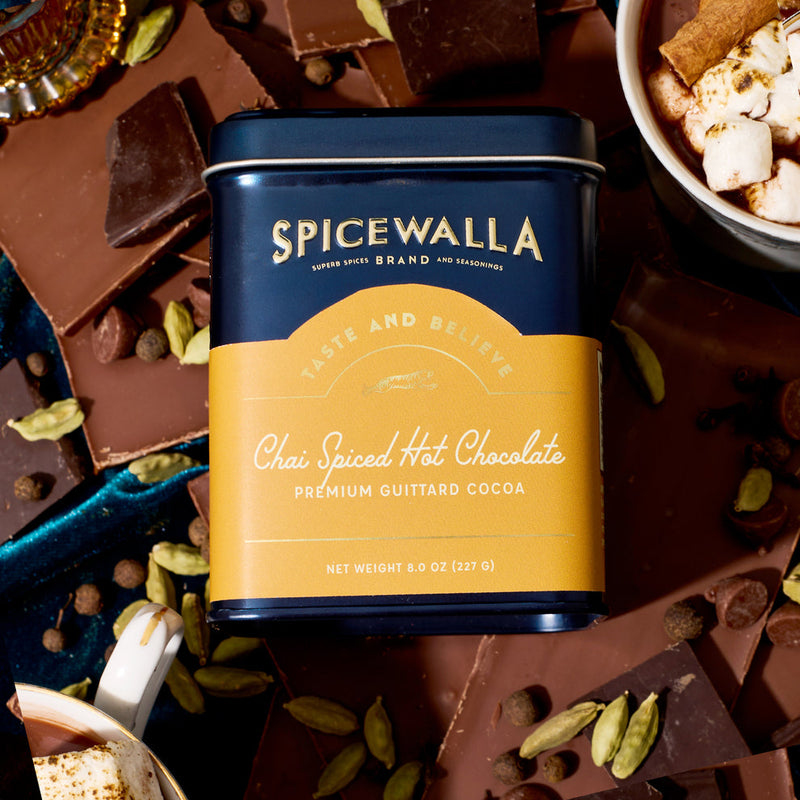Spicewalla | Chai Spiced Hot Chocolate tin surrounded by broken chocolate pieces and whole spices, with a cup of marshmallow-topped hot chocolate in the corner.