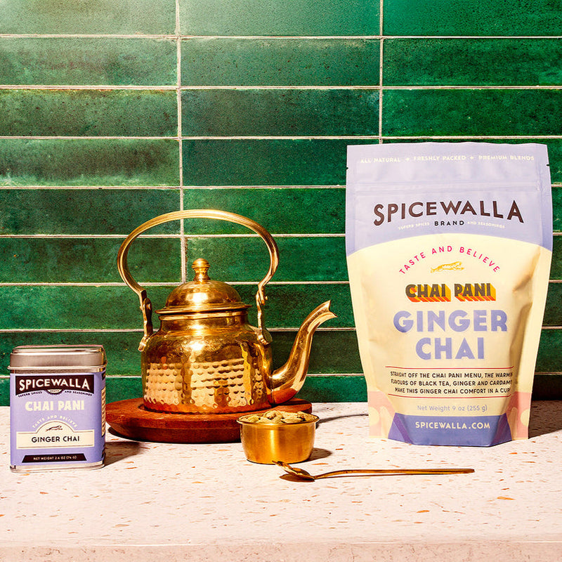 Spicewalla | Chai Pani Ginger Chai in tin and resealable bag, with a brass teapot, cup of cardamom pods, and teaspoon, on a speckled countertop.