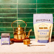 Spicewalla | Chai Pani Ginger Chai setup featuring a tin, brass kettle, cardamom bowl, spoon, and a large pouch on a countertop against green tiles.