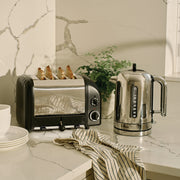 Dualit | 4 Slice Classic Toaster, Matt Black, on marble countertop with toasted bread, alongside a matching kettle, bowls, and a potted plant.