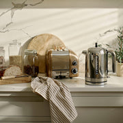 Dualit | 2 Slice Classic Toaster, Copper, with toast popping up, alongside artisan bread and glass jars, on a light kitchen countertop.