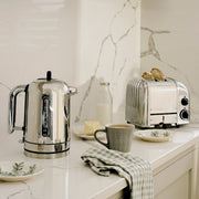 Dualit | 2 Slice Classic Toaster, Polished, with bread popping up on a marble countertop, surrounded by breakfast items like a kettle, mug, and butter dish.