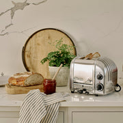 Dualit | 2 Slice Classic Toaster, Polished, with bread popping up, surrounded by rustic kitchen elements like crusty bread, jam jar, and a potted plant.
