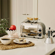 Dualit | 4 Slice Classic Toaster, Polished, on a marble countertop, with fruit tarts on the warming rack, alongside a moka pot and breakfast setup.