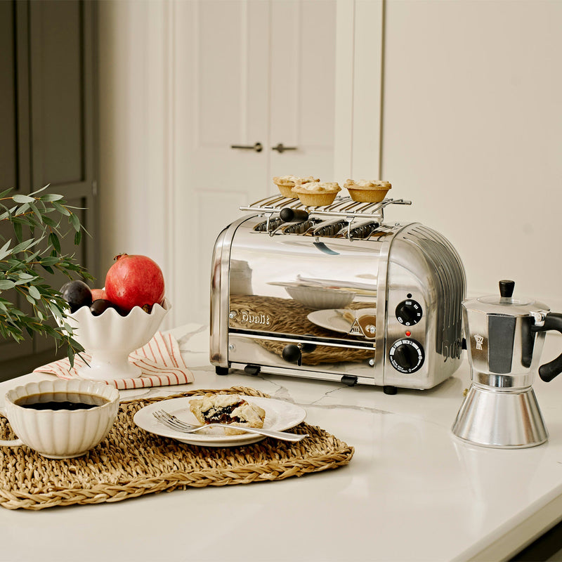 Dualit | 4 Slice Classic Toaster, Polished, on a marble countertop, with fruit tarts on the warming rack, alongside a moka pot and breakfast setup.