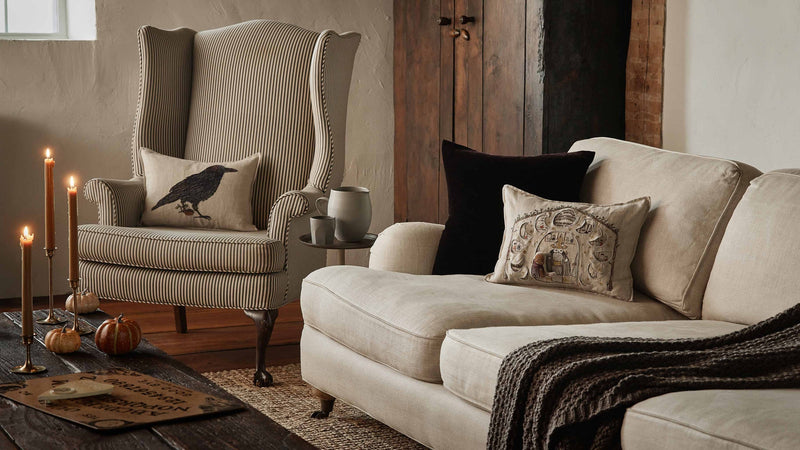Cozy living room corner with a Coral & Tusk Blackbird with Acorn Pillow on a beige pinstriped armchair, styled with autumn decor and rustic elements.