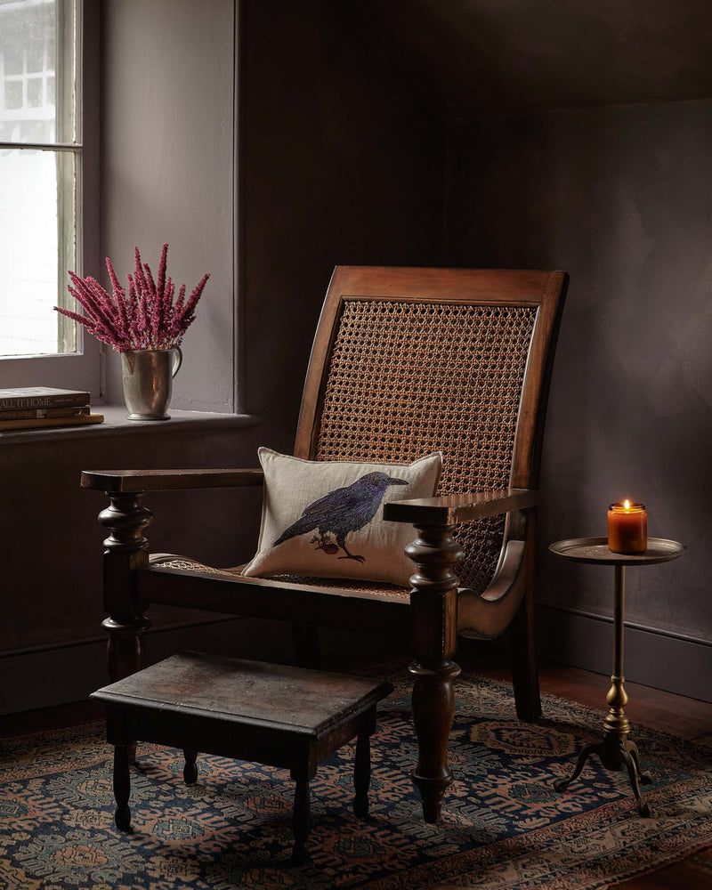 Coral & Tusk | Blackbird with Acorn Pillow: A linen throw pillow with an embroidered blackbird design, resting on a dark wooden armchair in a cozy room setting.