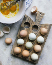 Farmhouse Pottery Crafted Wooden Egg Board displaying a dozen varied eggs, with a mixing bowl and whisk nearby, showcasing its kitchen utility.