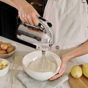 Close-up of Dualit Hand Mixer, Polished Chrome, blending batter in a white bowl, surrounded by baking ingredients like butter, eggs, and lemons, reflecting kitchen preparation.