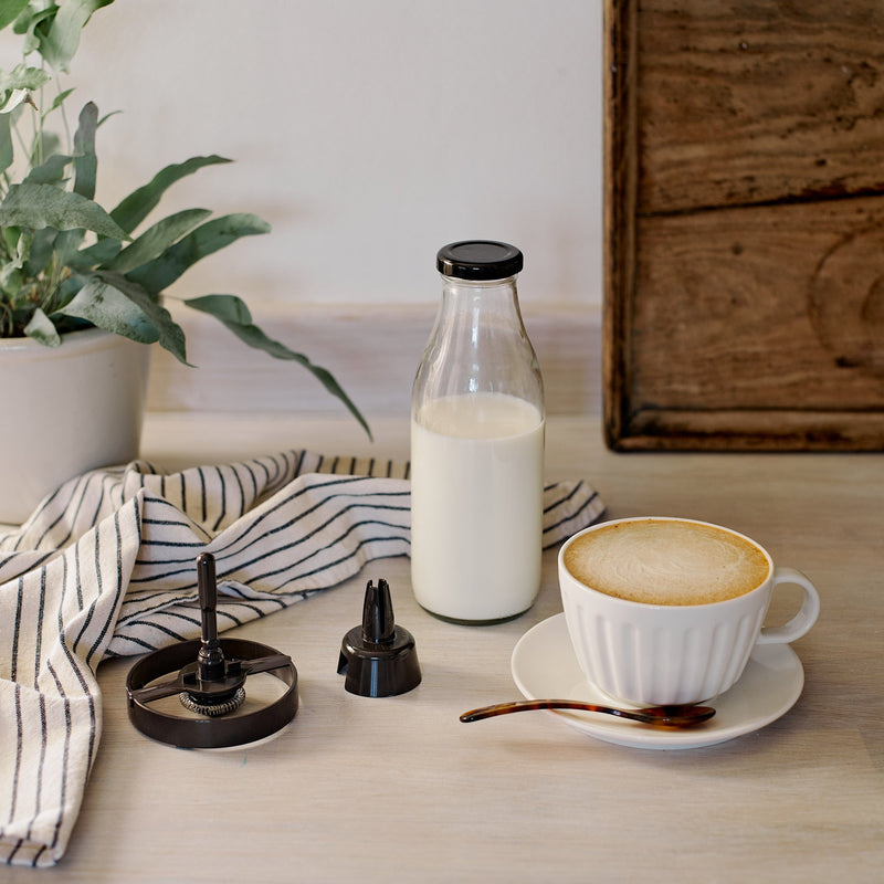 Dualit Hot Chocolate Maker and Milk Frother components on a tabletop, including a whisk attachment, near a ceramic cup of coffee, milk bottle, and striped towel.