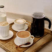 Dualit Hot Chocolate Maker and Milk Frother with three coffee cups, tortoiseshell spoon, and glass milk bottle on a rustic wooden tray.