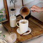Dualit Hot Chocolate Maker and Milk Frother pours frothy drink into mug; includes ceramic mug, saucer, spoon, glass milk bottle, and striped napkin on tray.