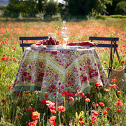 Caravan Home 71 x 71 Red & Green Tablecloth beautifully adorns an outdoor dining setup amidst a field of flowers, evoking French garden elegance.
