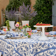 Granada Cornflower Blue Tablecloth by Caravan Home, 71 x 71, displayed on a party table with wine glasses, flowers, and a cake centerpiece.