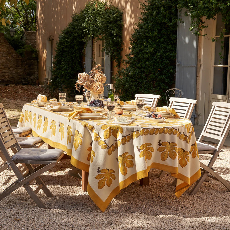 Caravan Home 59 x 86 Fig Citrine Tablecloth elegantly drapes an outdoor dining table set for a party, showcasing intricate handprinted details amidst a vibrant tablescape.