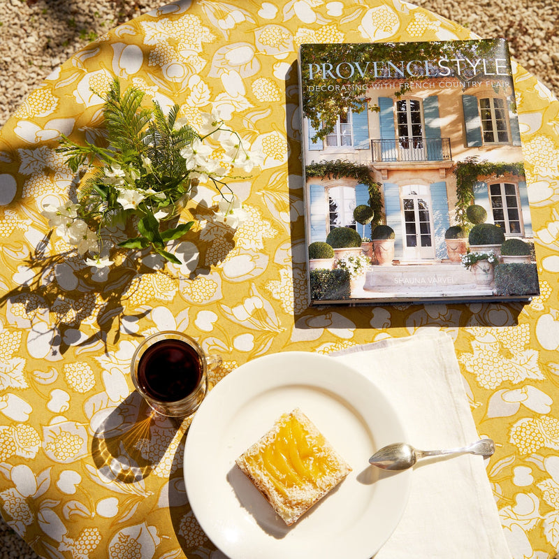 Forest Harvest Grey & Mustard Tablecloth on a table with a book, cake plate, spoon, and glass, embodying elegance and craftsmanship from Athens Cooks.