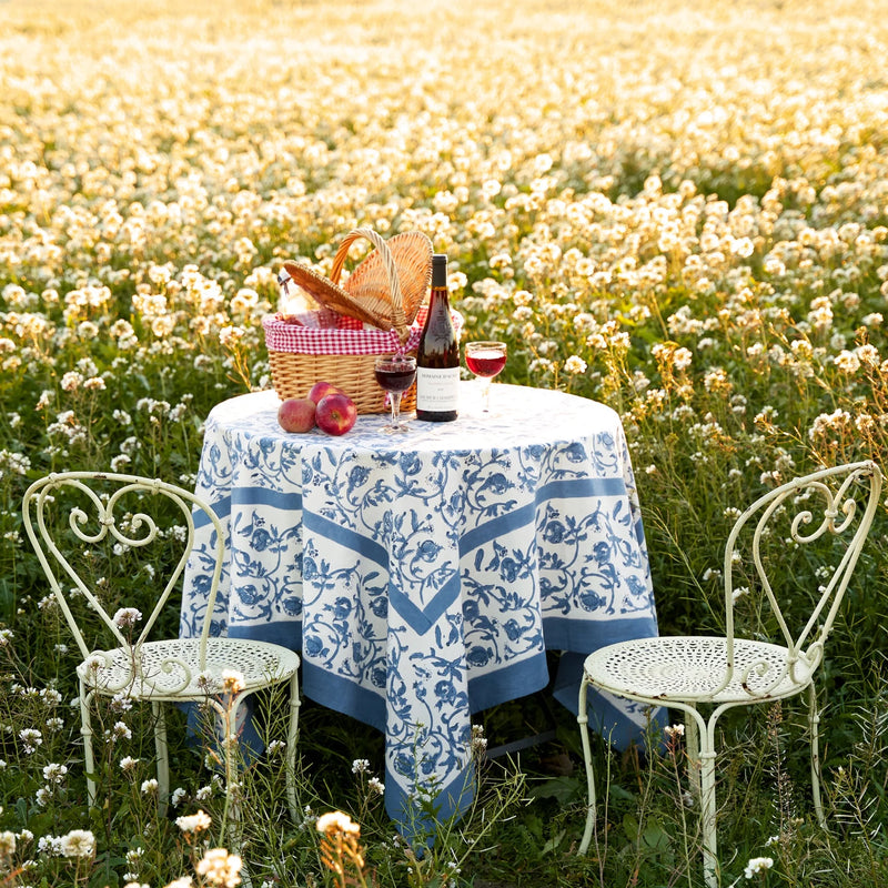 Granada Cornflower Blue Tablecloth adorning a picnic table amidst flowers, paired with wine and apples, highlighting its elegant French country style.