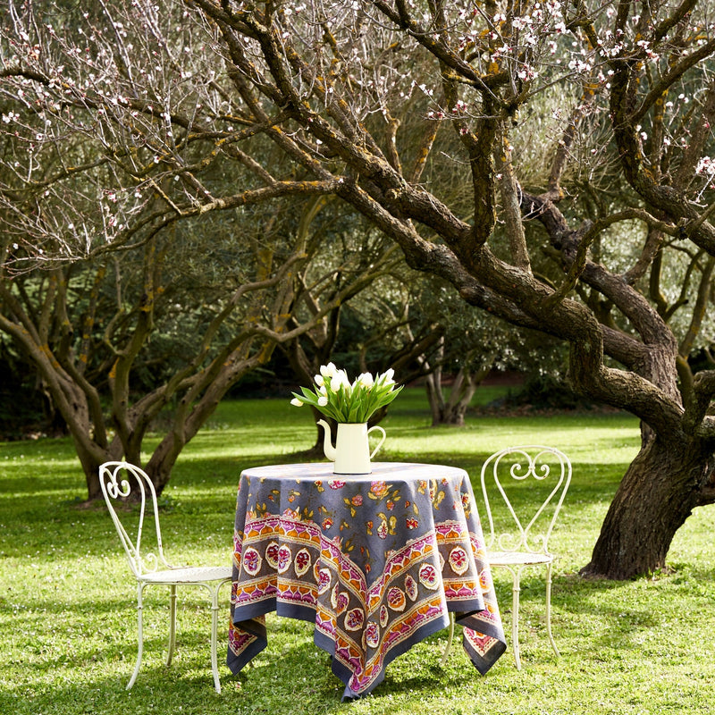 Caravan Home 71 x 71 Pansy Red & Grey Tablecloth on an outdoor table with white chairs, featuring handprinted floral patterns amidst a serene park setting.
