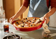 Person lifting a pizza from the Emile Henry | Chicago Deep Dish Pan, Charcoal, with kitchen towels, surrounded by dining essentials in a cozy setting.