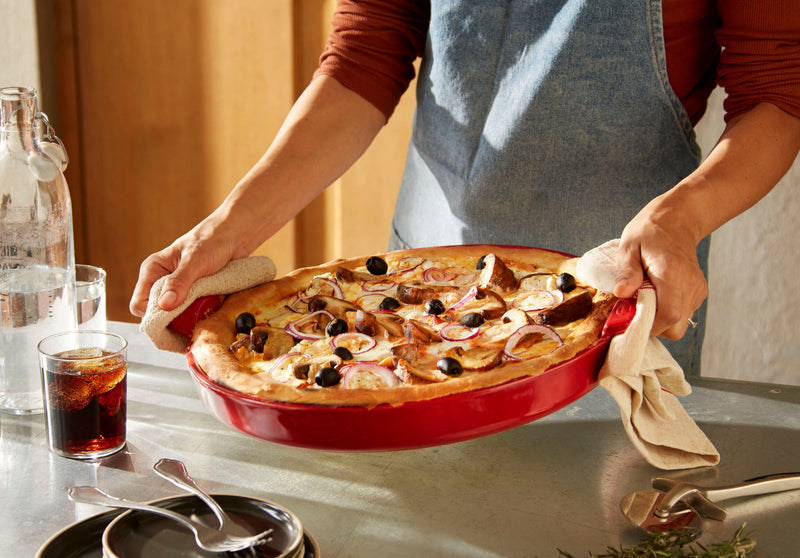 Person lifting a pizza from the Emile Henry | Chicago Deep Dish Pan, Charcoal, with kitchen towels, surrounded by dining essentials in a cozy setting.