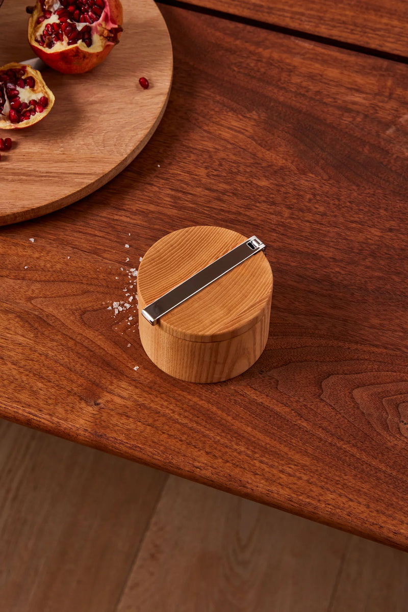 Holcomb Studio Stash Salt Cellar, a wooden container with a metal lid, displayed on a wooden surface alongside a cut pomegranate and a knife.