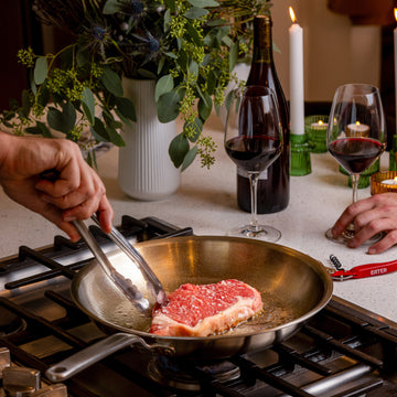 A steak sears in a Heritage Steel | Eater 5 Piece Essentials Set skillet, surrounded by wine, candles, and greenery, suggesting a cozy date-night atmosphere.