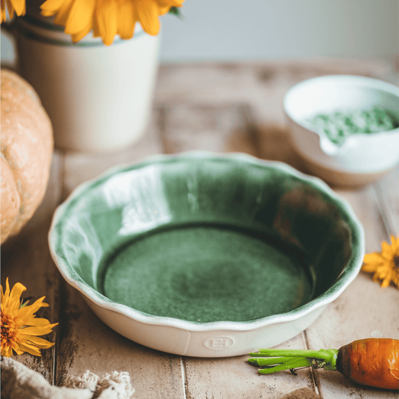 Emile Henry | 175th Anniversary Pie Dish, Green/Ivory, elegantly displayed with a carrot, green sauce, pumpkin, and yellow blooms on a wooden table.