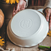 Emile Henry | 175th Anniversary Pie Dish, Green/Ivory, shown upside-down on a wooden table, held by hands, with sunflower bits and a pumpkin nearby.