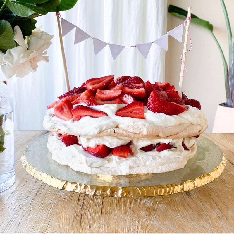 Annieglass Edgey Cake Stand elegantly displays a two-layered pavlova cake, highlighting its sophisticated glass design with 24k gold-edged detailing on a wooden table.