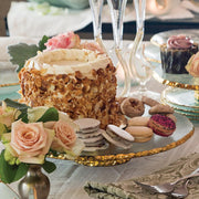 Annieglass Edgey Cake Stand elegantly displays a layer cake, macarons, and cookies, highlighting its gold-edged, glass design amidst a refined dessert spread.