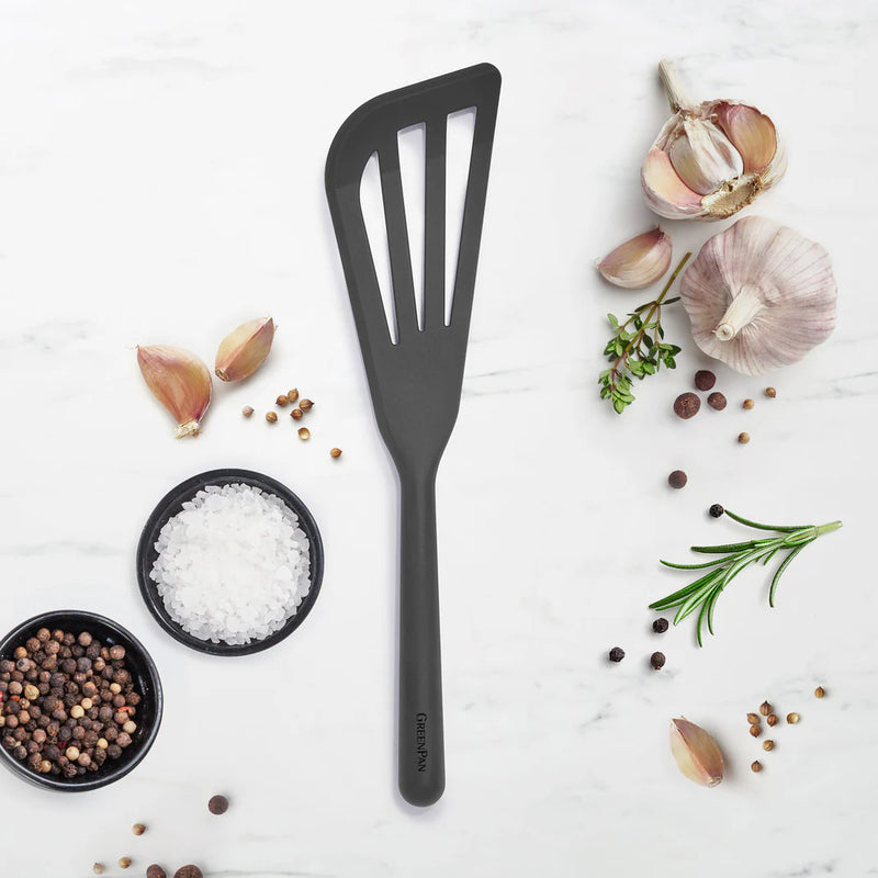GreenPan slotted cooking turner on marble, surrounded by bowls of sea salt, peppercorns, garlic, coriander seeds, and herbs, reflecting Athens Cooks' curated kitchen tool selection.