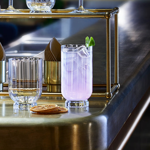 Close-up of Bormioli Florian Drinking Glasses, 14.5 oz., showcasing a fluted highball glass with a purple drink, beside an empty glass on a brass bar counter.