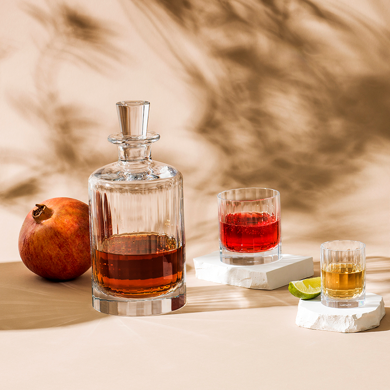 Richard Brendon | Fluted Large Decanter with ribbed design, partially filled with amber liquid, alongside pomegranate and two glasses on stone blocks with beverages.