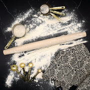 J.K. Adams French Tapered Rolling Pin rests on a floured counter, surrounded by brass measuring tools, highlighting its ergonomic design and textural grip for baking enthusiasts.