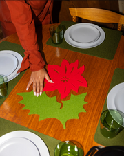 Graf Lantz Merino Wool 10 Poinsettia Trivet - Red on a table, featuring a hand and festive setting, ideal for protecting surfaces during holiday meals.